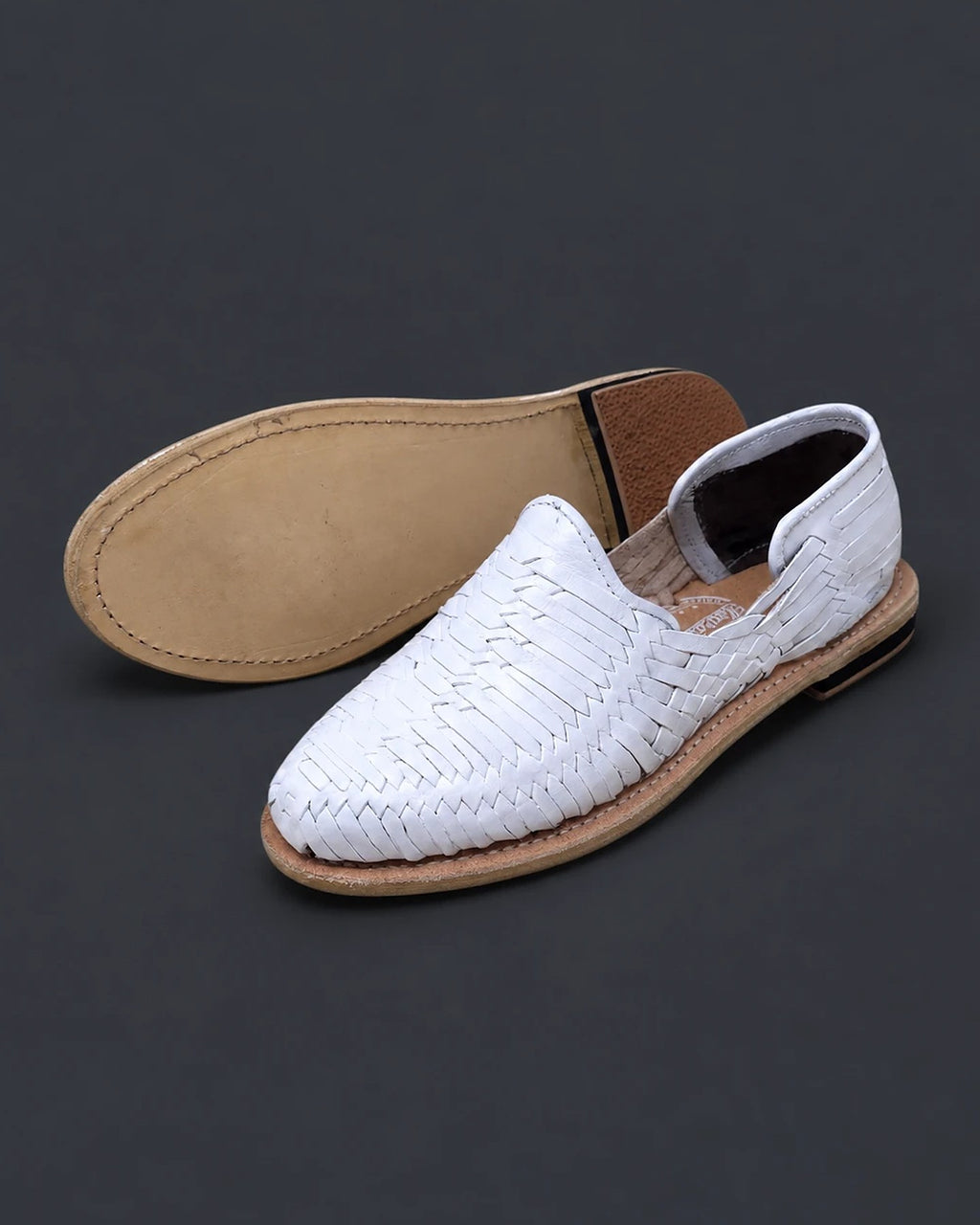 Natural leather sole stitched around the edge with a low stacked heel, beside a women's white closed toe huarache sandal with a stamped leather insole