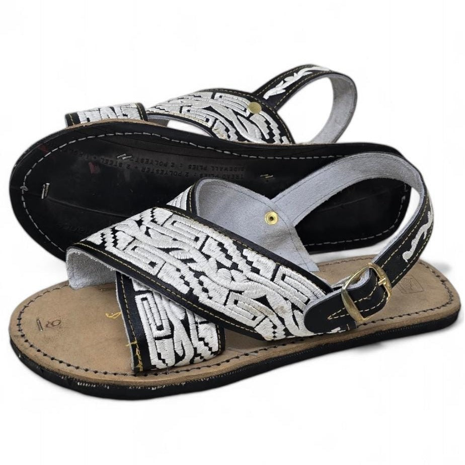 Black and white men's huarache sandals, one turned over to show the tire sole with its lettering