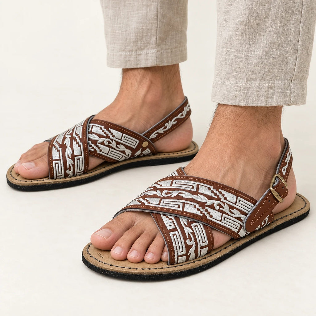 Men's embroidered huarache sandals in brown and white with cross straps and a buckled back strap, worn with linen pants